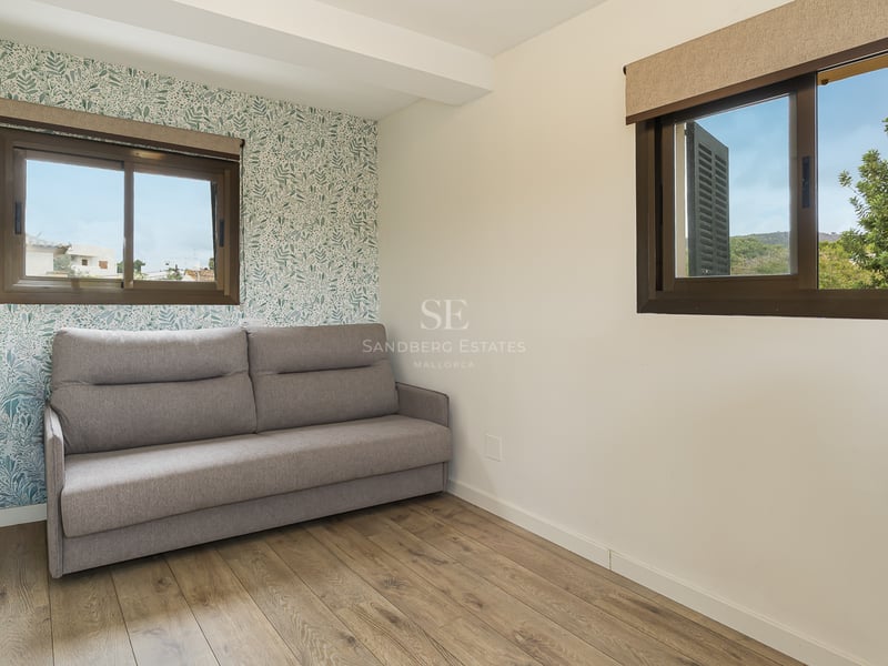 Chambre moderne avec un canapé gris, parquet en bois, papier peint à motif végétal et deux fenêtres.