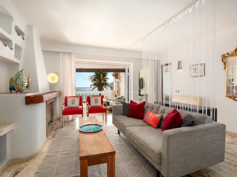 Salon avec canapé gris, chaises rouges, cheminée rustique et grandes baies vitrées ouvrant sur la mer et une terrasse.