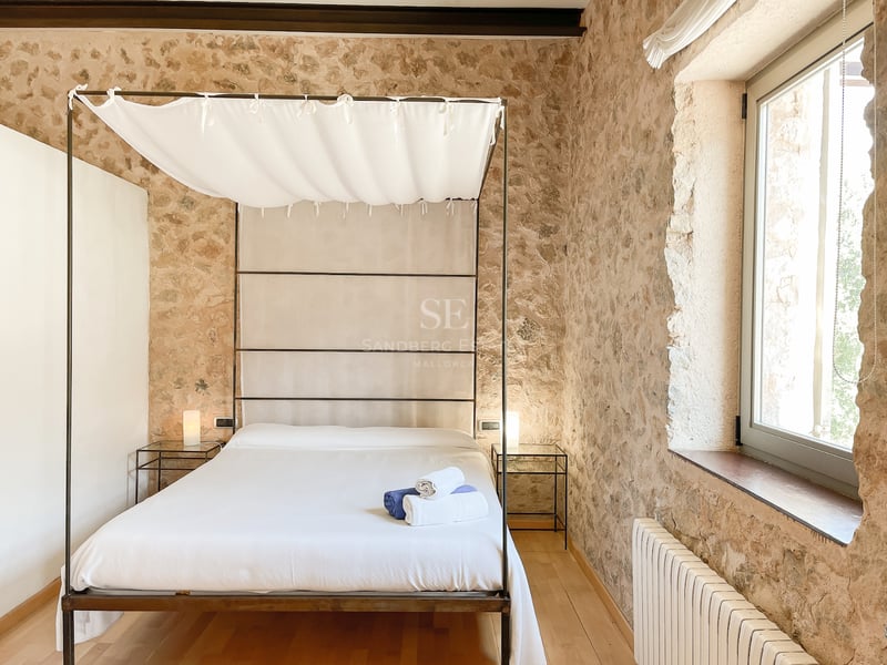 Lit à baldaquin avec draps blancs devant un mur en pierre naturelle dans une chambre lumineuse avec parquet clair.