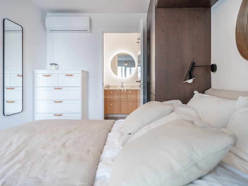 Chambre à coucher avec lit blanc ouvrant sur une salle de bain avec miroir rond rétroéclairé et meuble en bois.