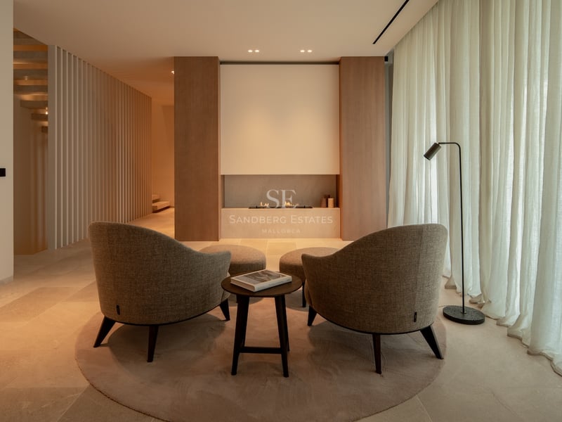 Salon minimaliste avec deux fauteuils beiges face à une cheminée linéaire, éclairage chaleureux et boiseries.