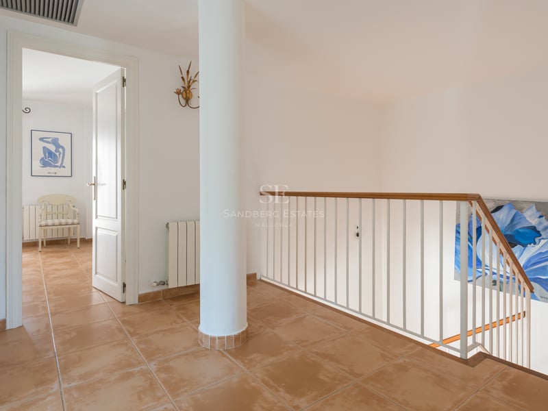 Couloir avec carrelage en terre cuite, pilier blanc et rampe d'escalier menant à une porte ouverte.