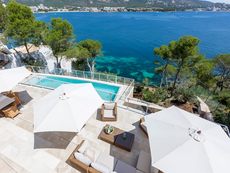 Vue en plongée d'une terrasse en pierre avec parasols blancs, mobilier de salon et piscine surplombant la mer.
