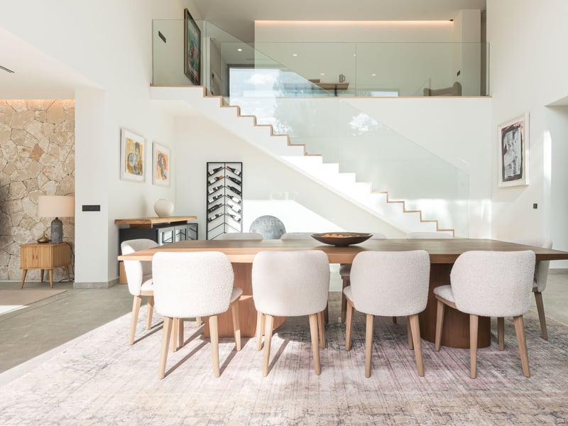 Salle à manger minimaliste avec table en bois, chaises blanches en bouclé et escalier en verre moderne.
