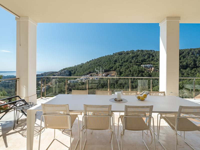 Terrasse couverte moderne avec table blanche et garde-corps en verre, vue sur les collines verdoyantes et la mer.