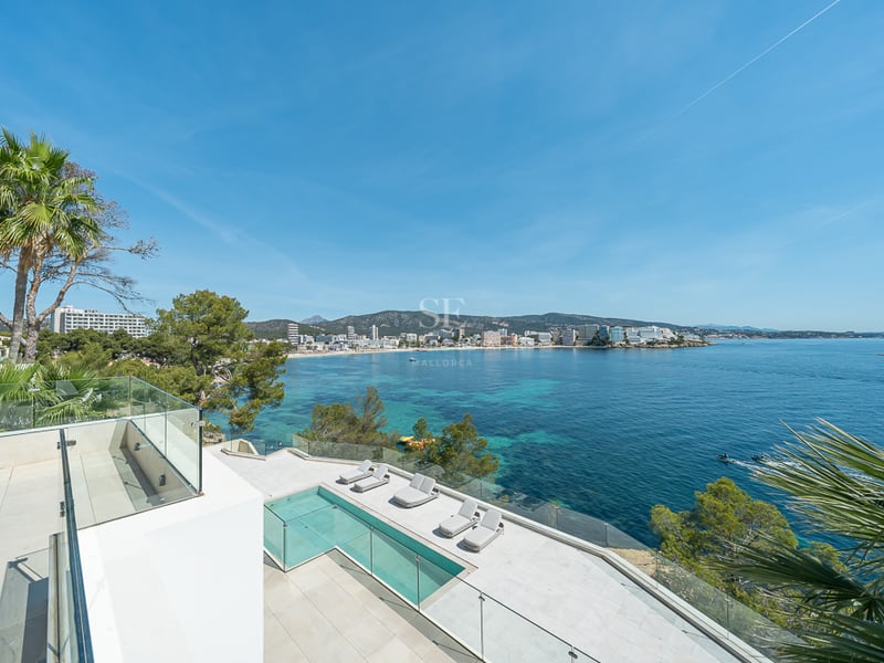 Terrasse blanche moderne avec piscine à débordement et garde-corps en verre surplombant une baie turquoise.