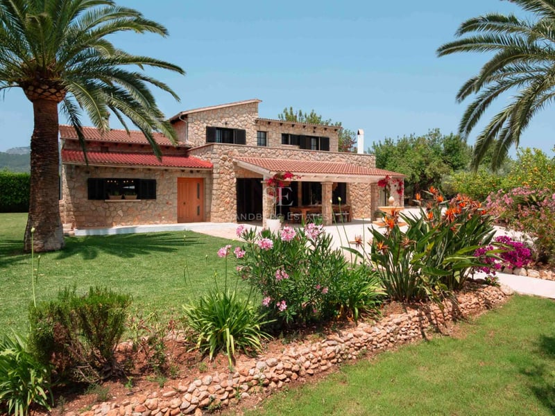 Villa en pierre naturelle de deux étages avec toits en terre cuite, palmiers et fleurs éclatantes par temps clair.