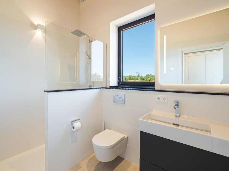 Baño minimalista blanco con ducha de cristal, inodoro suspendido y mueble de lavabo oscuro bajo una ventana.