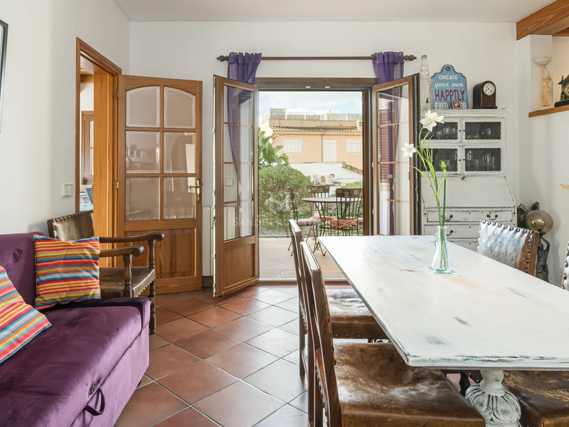 Salle à manger avec sol en terre cuite, table en bois blanc vieilli et portes ouvertes sur un patio ensoleillé.