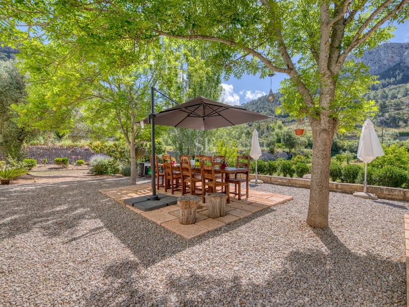 Table et chaises en bois sous un parasol sur une terrasse en gravier entourée d'arbres et de montagnes.