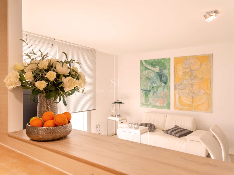 Salon lumineux avec canapé en cuir blanc, peintures abstraites et comptoir en bois avec fleurs.