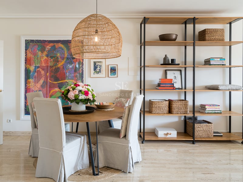 Salle à manger avec table ronde en bois, chaises beiges, suspension en rotin et étagère moderne sur sol en marbre.