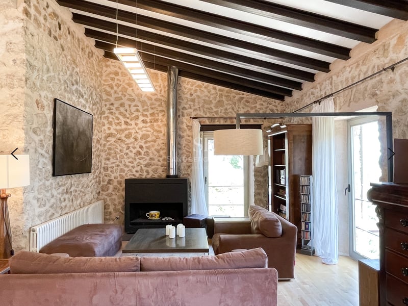 Salon avec murs en pierre, plafond à poutres sombres, cheminée moderne et grandes fenêtres avec lumière naturelle.