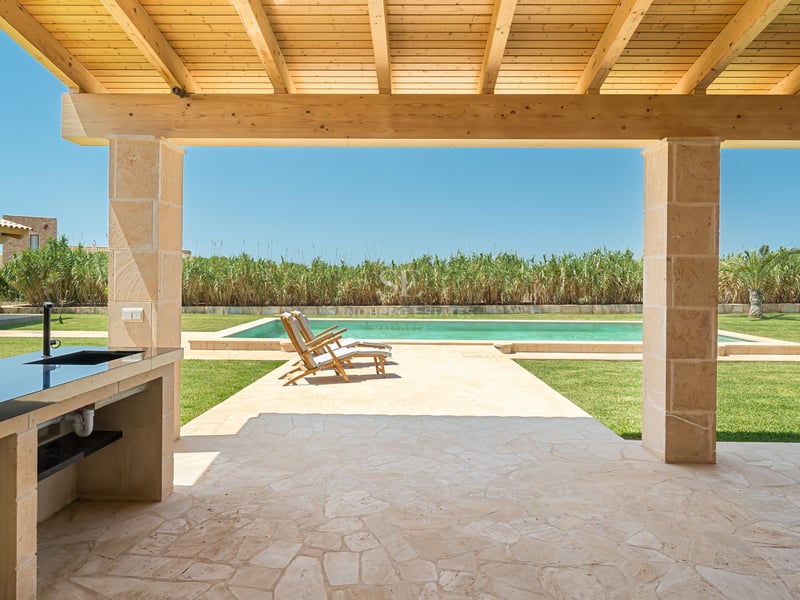 Porche de piedra con cocina exterior mirando hacia una piscina y jardín bajo un cielo despejado.