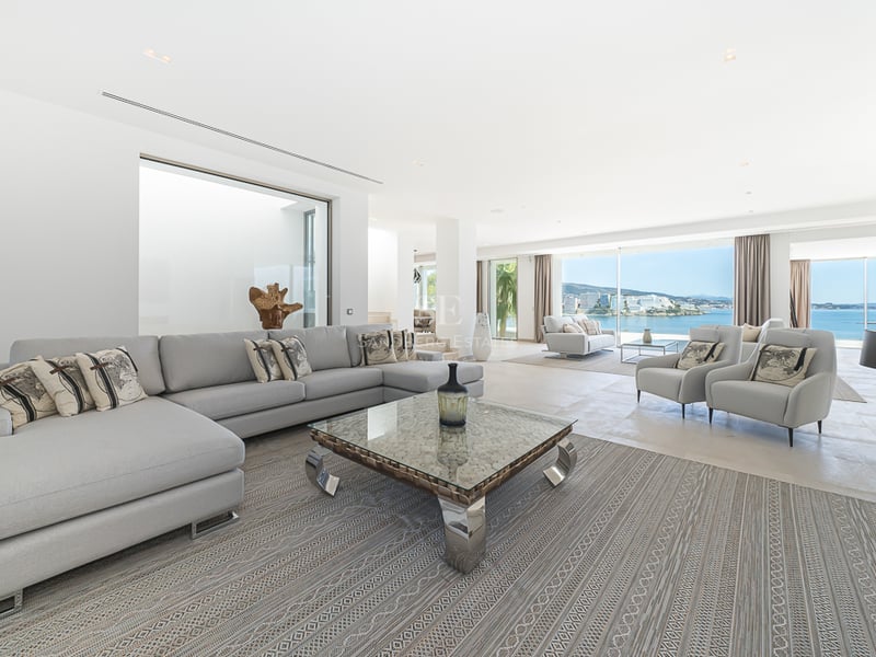 Grand salon moderne avec canapés gris clair, table basse en verre et grandes baies vitrées donnant sur la mer.