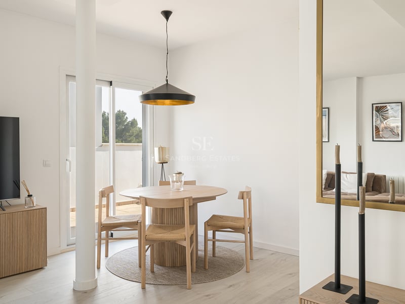 Salle à manger lumineuse avec table ronde en bois, chaises en osier, lampe noire et accès à la terrasse.