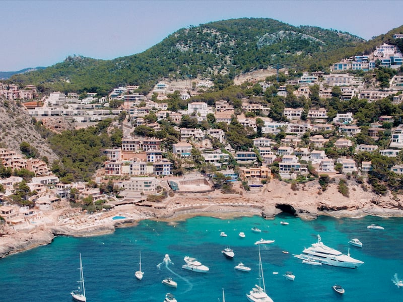 Vue en plongée de maisons blanches sur une colline verdoyante surplombant une baie avec des yachts.