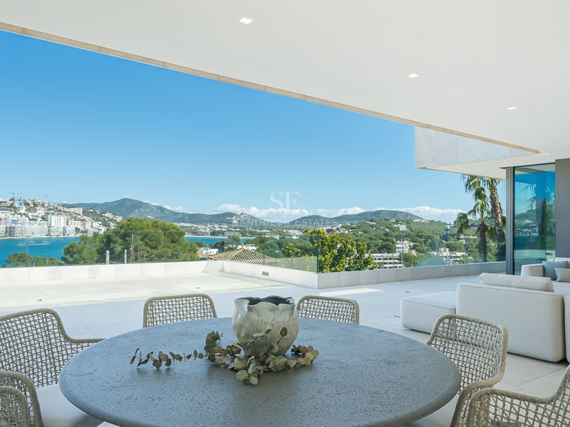Terrasse moderne avec canapés blancs, table en pierre et garde-corps en verre surplombant la mer et les montagnes.