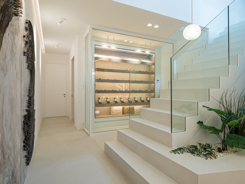 Modern white staircase with glass railing next to a glass-enclosed illuminated wine display case.