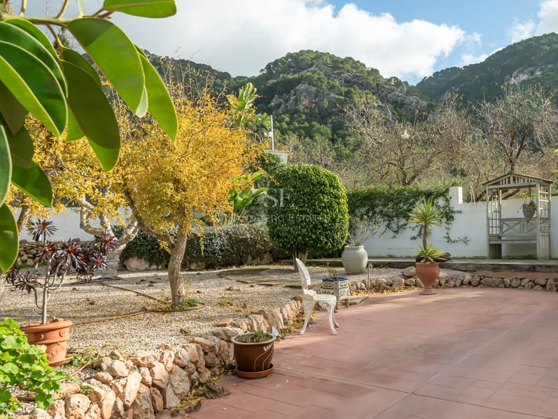 Terrasse en terre cuite et jardin avec murs en pierre et arbres matures sur fond de montagnes rocheuses.
