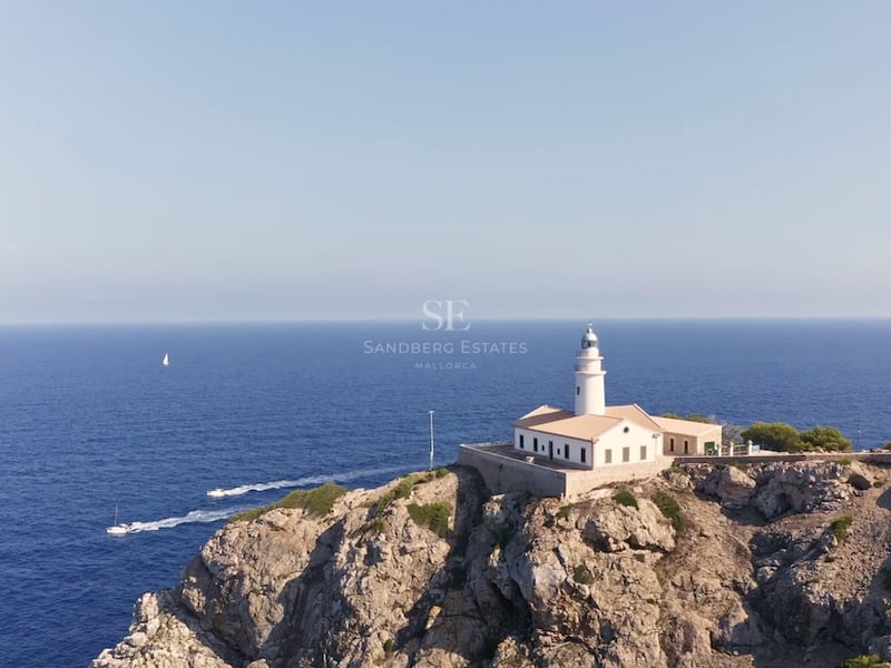 Aerial view of a white lighthouse perched on rugged cliffs overlooking the deep blue Mediterranean Sea with sailboats.