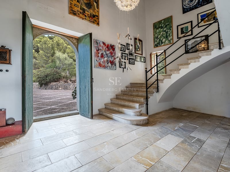 Vaste hall d'entrée avec sols en pierre, hauts plafonds, escalier en pierre et grandes portes vertes ouvertes.