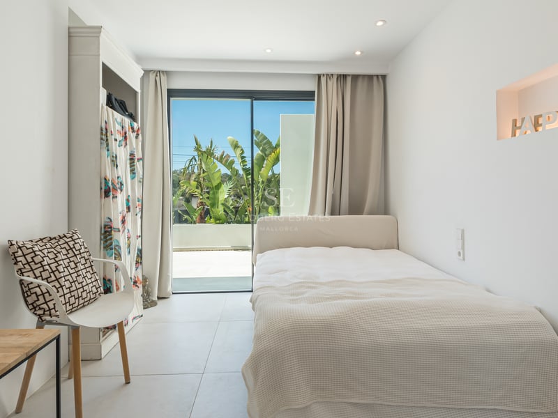 Chambre moderne blanche avec lit simple, grandes baies vitrées donnant sur une terrasse et végétation tropicale.
