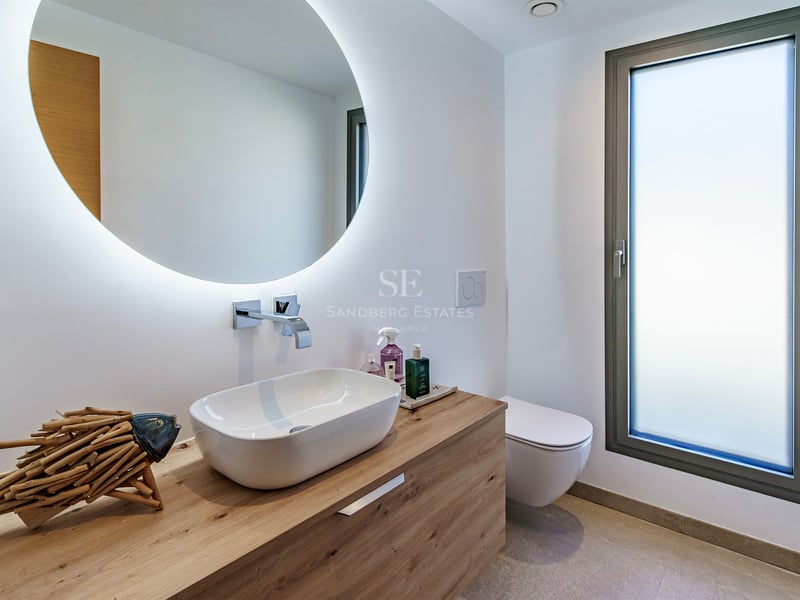 Salle de bain moderne avec vasque sur meuble en bois, miroir rond éclairé et fenêtre en verre dépoli.