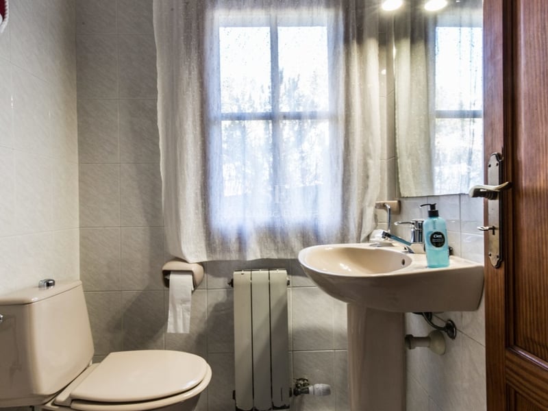Petite salle de bain avec toilettes et lavabo colonne beige, fenêtre avec rideau et murs carrelés clairs.