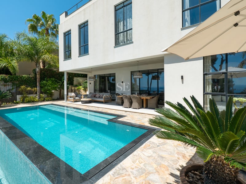 Scène extérieure lumineuse avec piscine turquoise, terrasse couverte et villa blanche moderne entourée de palmiers.