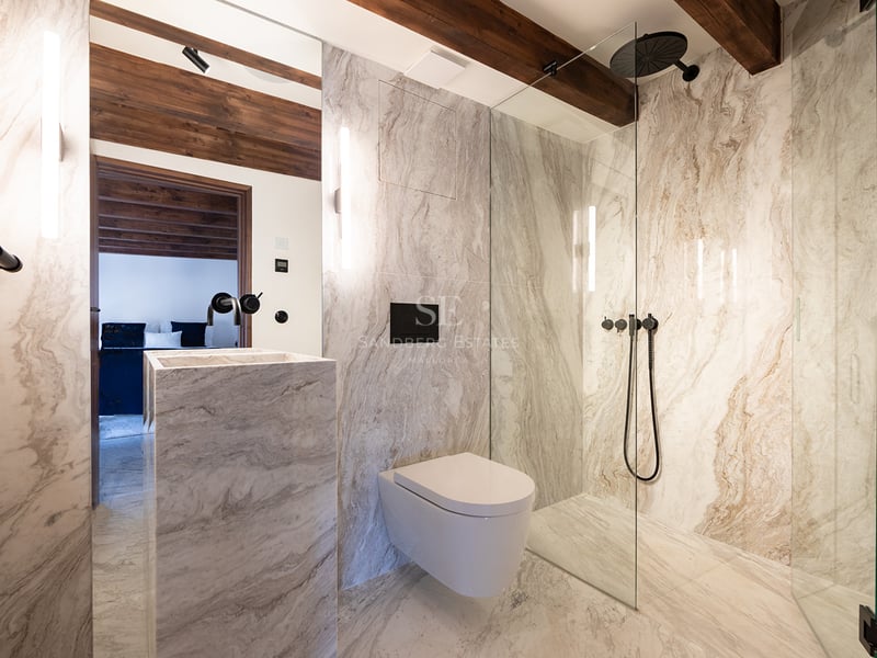 Baño moderno con paredes de mármol, ducha de cristal, grifería negra mate y vigas de madera vista en el techo.