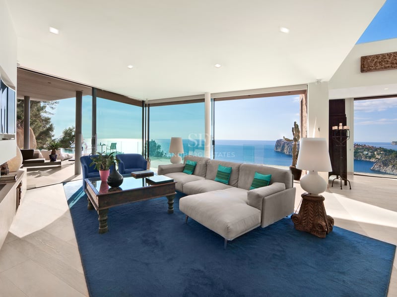 Salon moderne avec canapé gris, tapis bleu et grandes baies vitrées offrant une vue imprenable sur l'océan.