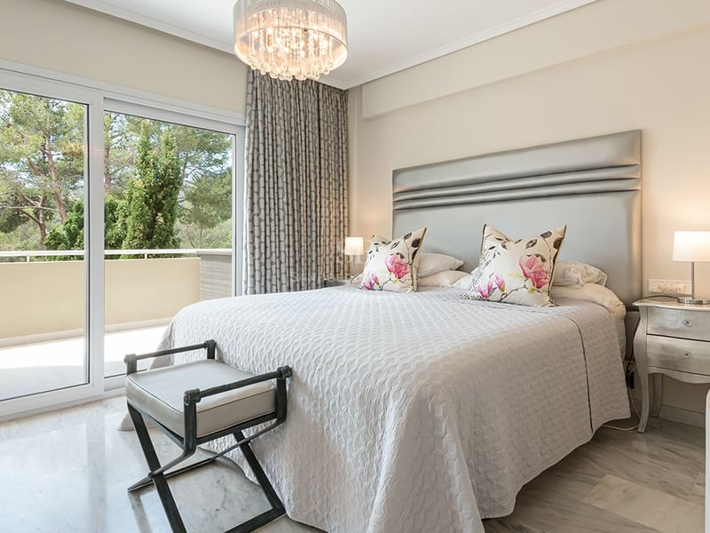Chambre principale lumineuse avec sol en marbre, tête de lit argentée et accès terrasse vue sur les arbres.