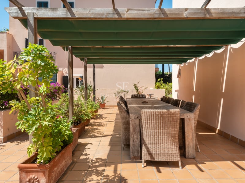 Espace repas extérieur sur une terrasse en terre cuite avec pergola en bois, store vert et chaises en osier.