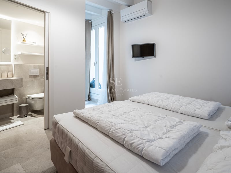 Chambre lumineuse avec literie blanche et porte ouverte sur une salle de bain moderne en pierre.