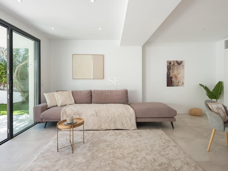 Salon moderne avec canapé mauve, tapis moelleux et baies vitrées s'ouvrant sur un jardin verdoyant.