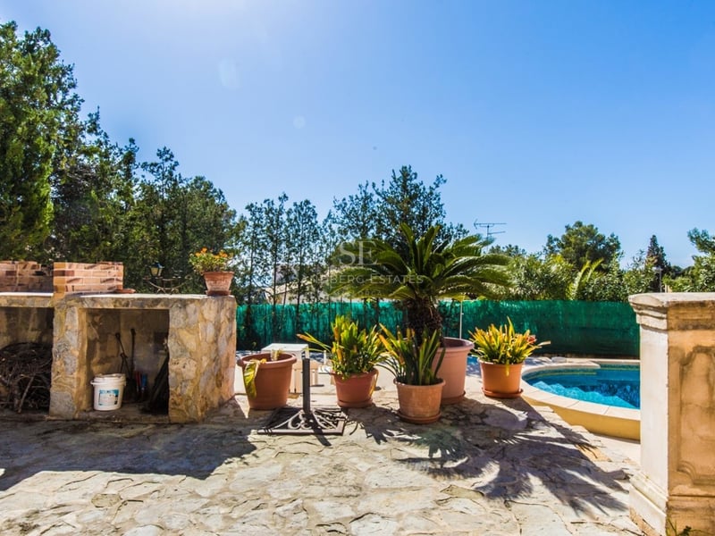 Terrasse en pierre ensoleillée avec piscine bleue, barbecue intégré et plantes en pots sous un ciel dégagé.