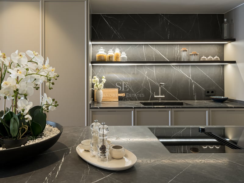 Modern kitchen featuring dark marble countertops, integrated appliances, and warm LED shelf lighting with white orchids.