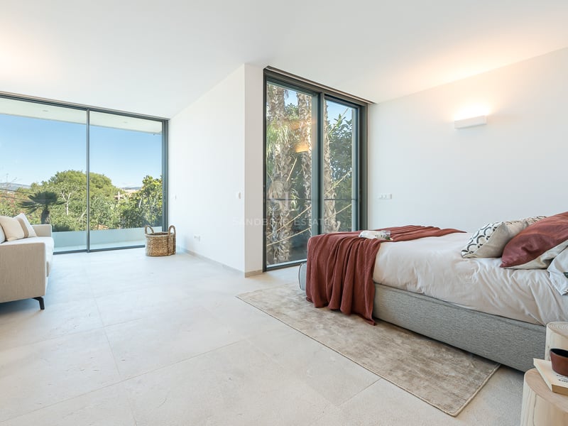 Chambre lumineuse avec un grand lit, un canapé beige et des baies vitrées donnant sur un jardin de palmiers.