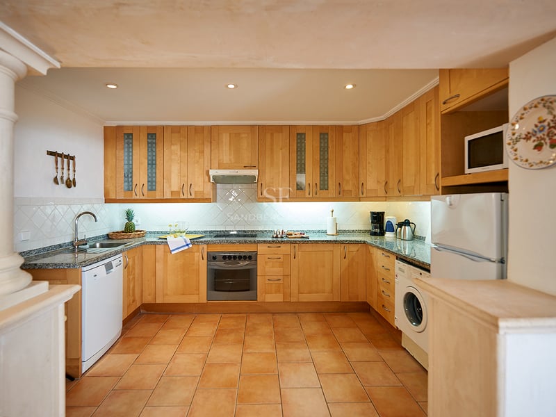 Cuisine rustique avec armoires en bois clair, plans de travail en granit, sol en terre cuite et électroménager.