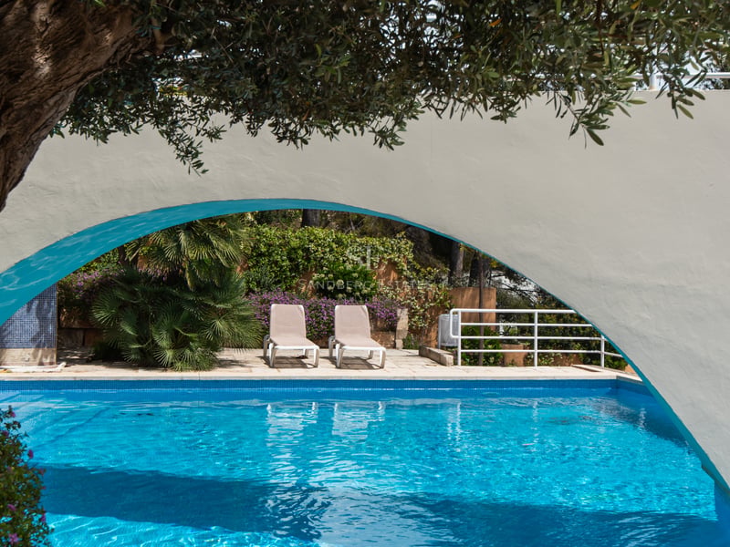 Piscine bleue et transats encadrés par une arche blanche et une végétation luxuriante dans un jardin.