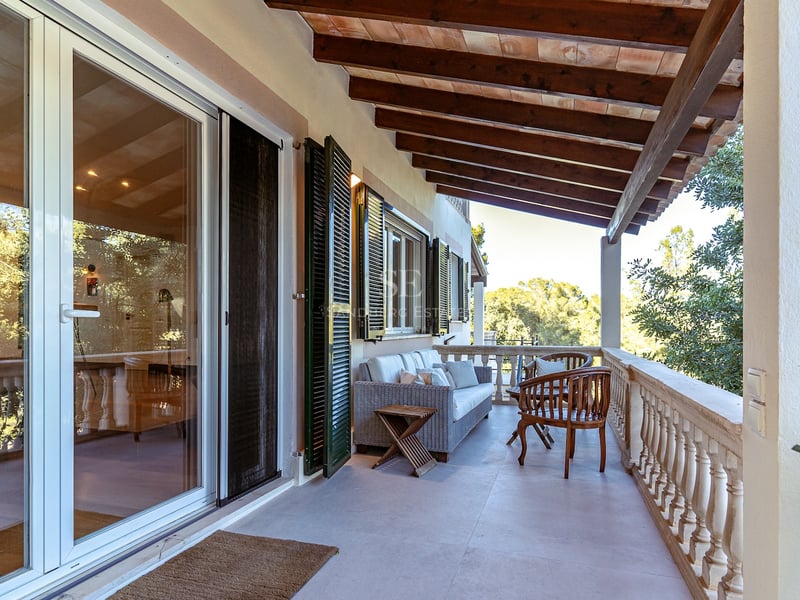 Terrasse couverte avec poutres apparentes, canapé en osier, balustrade en pierre et vue sur la verdure.