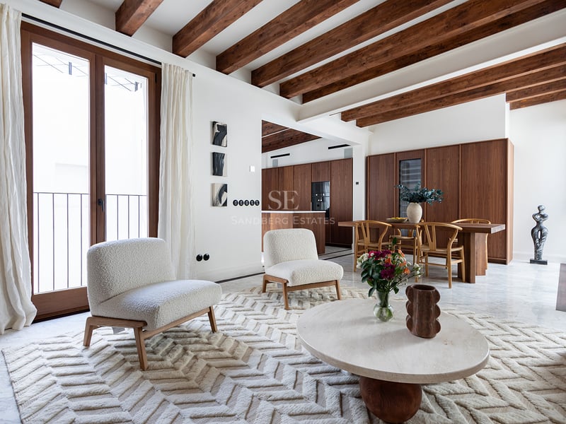 Sala de estar moderna con vigas de madera oscura, sillones bouclé blancos, suelo de mármol y comedor al fondo.