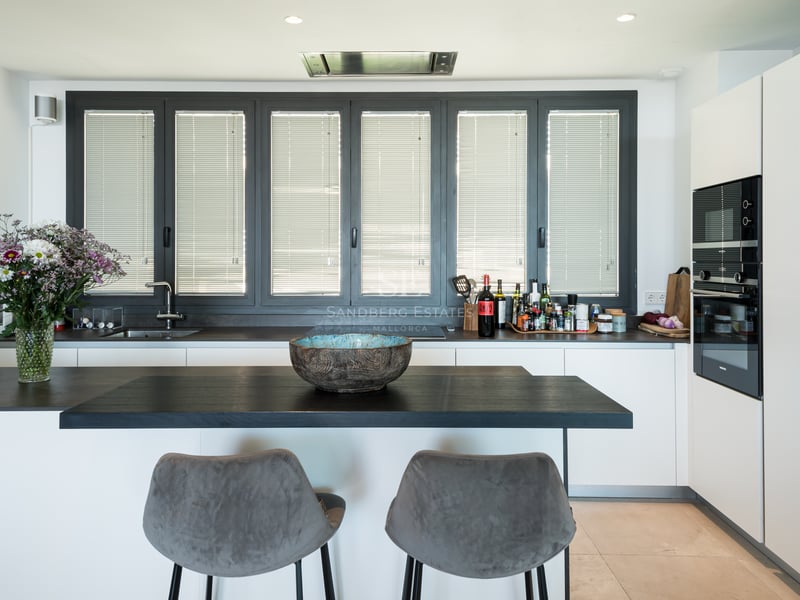 Cuisine blanche moderne avec plans de travail sombres, îlot avec deux tabourets gris et grandes fenêtres.