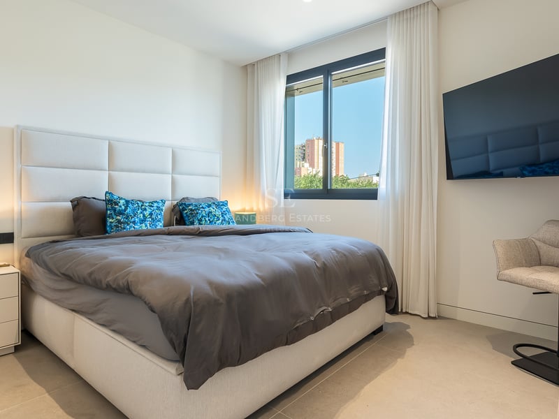 Chambre moderne avec grand lit, tête de lit rembourrée grise, TV murale et fenêtre lumineuse.