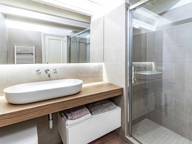Salle de bains avec vasque ovale sur plan de travail en bois, douche vitrée et poutres apparentes blanches.