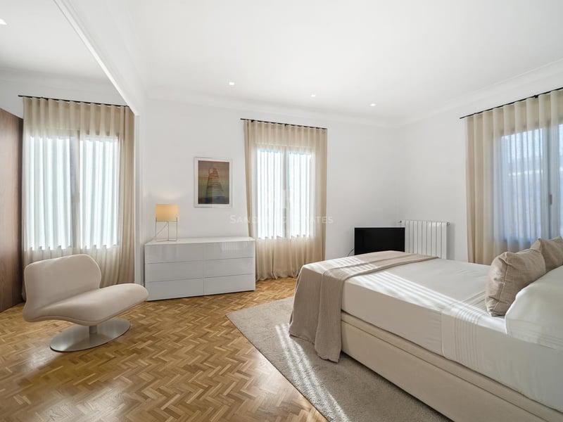 Chambre lumineuse avec parquet en chevrons, lit double, fauteuil design et rideaux voilages.