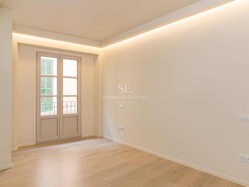 Empty modern bedroom with light oak flooring, cream walls, cove lighting, and a glass door to a Juliet balcony.