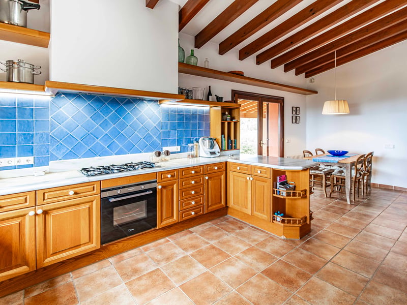 Cuisine rustique avec meubles en bois, crédence bleue, sol en terre cuite et plafond à poutres apparentes.