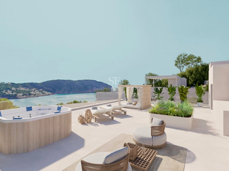 Terrasse ensoleillée avec jacuzzi en bois, transats et fauteuils surplombant une baie côtière.